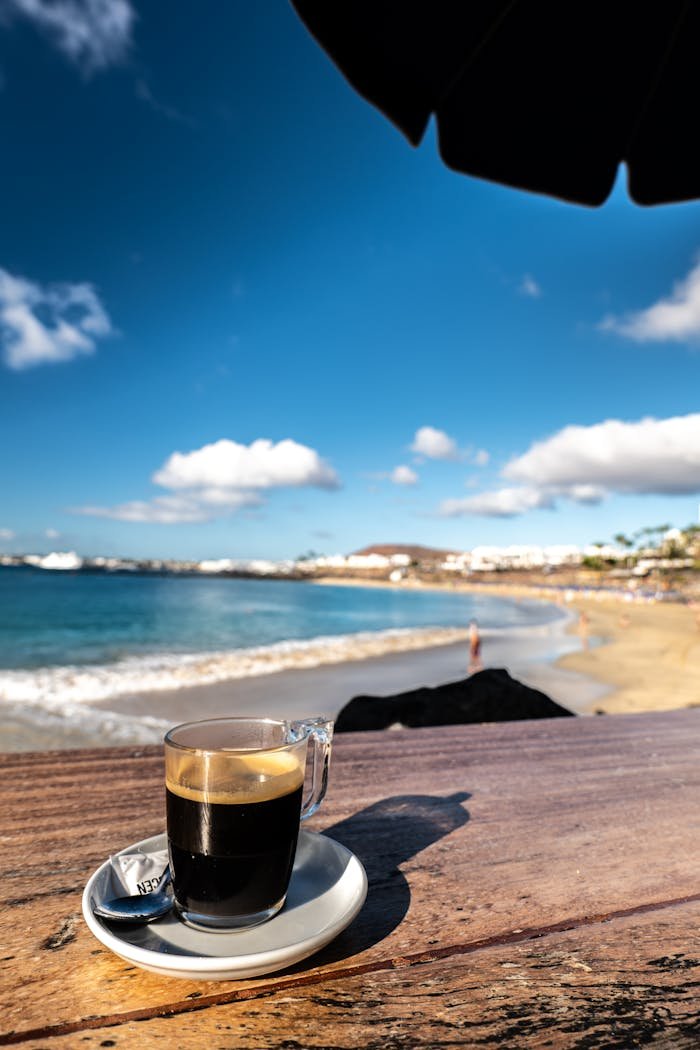 Sunlit coffee cup on a wooden table by Lanzarote's sandy beach, perfect for a summer vacation vibe.